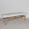 angled view of a large creamy white cowhide bench seat on a teak wood base.