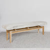 angled view of an off-white cowhide bench seat made from a single cowhide and secured to a teak wood base, on a grey large-looped rug