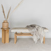 front view of the cream and caramel colored hide used on a Corcovado hair on hide bench seat with a wool throw blanket on top of the bench and a small wooden side table and woven rattan basket next to the bench