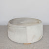 extra large round hair on hide ottoman coffee table with leather stitching and small teak wood feet on a grey and white rug