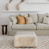 square neutral colored hair on hide ottoman coffee table in a family room with a large earthy colored sofa and wooden side table