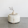 cream colored light round cowhide ottoman coffee table with a rattan tray and vase on top