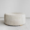 Side Angle view of Corcovado Designs round hide ottoman coffee table with a grey stripe and leather stitching placed on a white looped floor rug